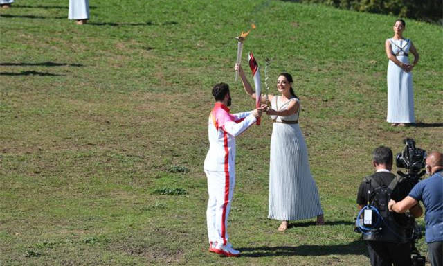 10月18日，扮演最高女祭司的希腊演员桑西·乔治乌（中）点燃第一棒火炬手、希腊高山滑雪运动员扬尼斯·安东尼乌的火炬。