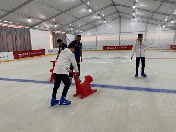 一名初学者在冰上体验区扶着滑板助手缓慢滑行，防止摔跤。旁边有教练在辅导。